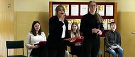 Na korytarzu szkolnym pod ścianą znajduje się stojąca gazetka, na której powieszone zostały plakaty informacyjne na temat depresji, przed gazetką siedzi na krzesłach 4 uczniów klasy 8, którzy będą prezentować temat zgromadzonym widzom z pozostałych klas, na środku stoją dwie uczennice, które wykonują piosenkę do mikrofonu