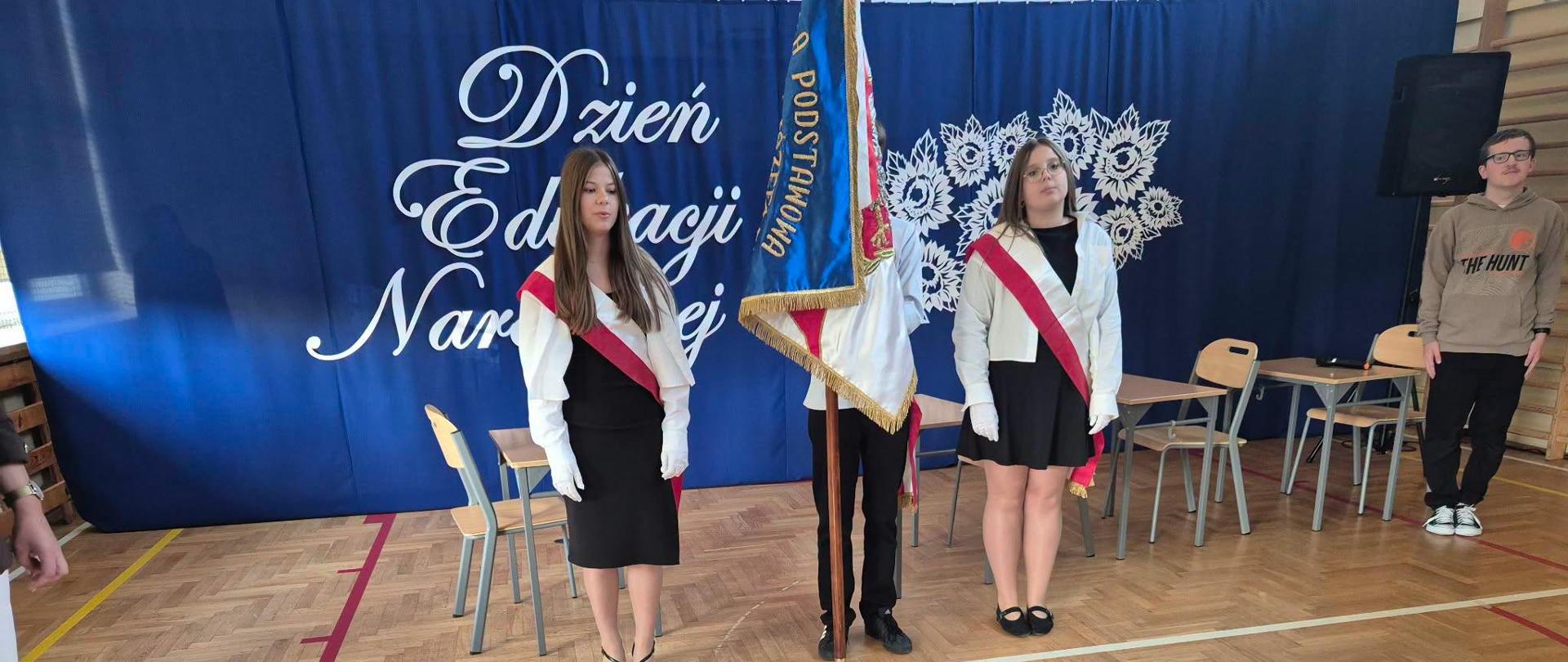 Na zdjęciu przedstawiono uroczystość szkolną z okazji Dnia Edukacji Narodowej, odbywającą się na sali gimnastycznej. Na tle granatowej dekoracji z napisem „Dzień Edukacji Narodowej” stoją uczniowie w strojach galowych, z biało-czerwonymi szarfami i białymi rękawiczkami. W centrum znajduje się poczet sztandarowy, który trzyma sztandar szkoły. W tle widać szkolne ławki i krzesła, a na podłodze – oznaczenia boiska.
Fotografia ukazuje podniosły, uroczysty charakter wydarzenia, podkreślając szacunek dla tradycji szkolnych, patriotyzm oraz znaczenie edukacji i pracy nauczycieli. Jest to chwila, która łączy społeczność szkolną we wspólnym świętowaniu.
