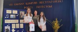 Na scenie podczas „VIII Gminnego Konkursu Recytatorskiego – Poeci Dzieciom” stoją trzy osoby: dwie uczennice trzymające dyplomy oraz nauczycielka. Obok nich znajduje się tablica poświęcona Mieczysławowi Foggowi, a w tle widoczna jest dekoracja z napisem konkursu i wazon z żółtymi kwiatami.