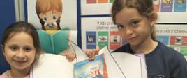 Dwie dziewczynki trzymają w ręku wykonaną z papieru otwartą książkę z napisem "Ogólnopolski Dzień Głośnego Czytania" oraz książką Janiny Porazińskiej "Szewczyk Dratewka"