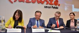 Cztery osoby siedzą przy stole i podpisują dokumenty. Za nimi znajduje się ściana z napisem „Mazowsze. serce Polski” w czerwono-czarnych literach. Przed każdą osobą ustawiono tabliczki z imionami i nazwiskami – od lewej: Janina Ewa Orzełowska, Adam Struzik, Wójt Gminy Pokrzywnica Adam Dariusz Rachuba oraz Skarbnik Gminy – Pani Magdalena Lusa. Na stole stoją butelki z wodą.