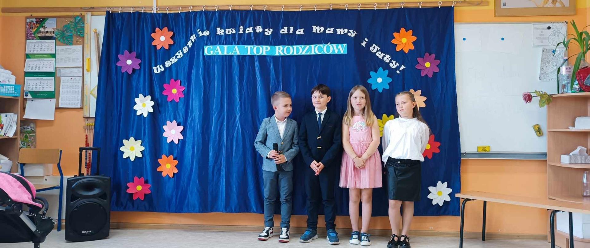 Dwoje chłopców w marynarkach z mikrofonami w rękach. Obok nich dwie dziewczynki. Na ścianie napis gala top rodziców