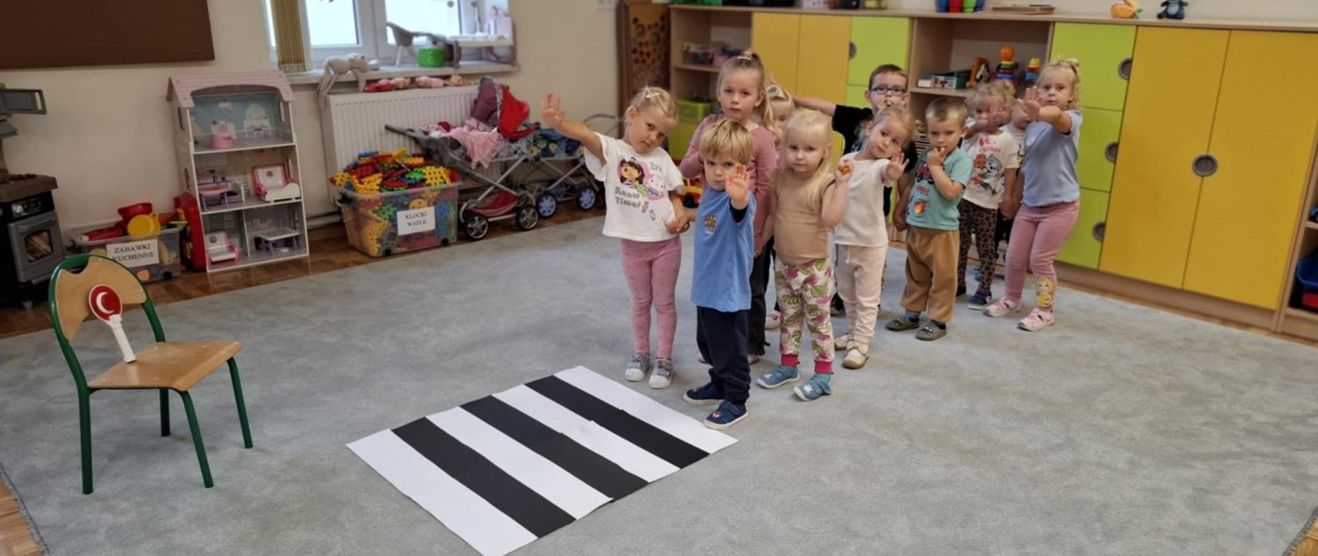 Grupa dzieci ustawionych w pary stoi na dywanie na którym rozłożona jest mata z pasami drogowymi. Po obu stronach maty ustawione są samochody tak by całość przypominała prawdziwą drogę.