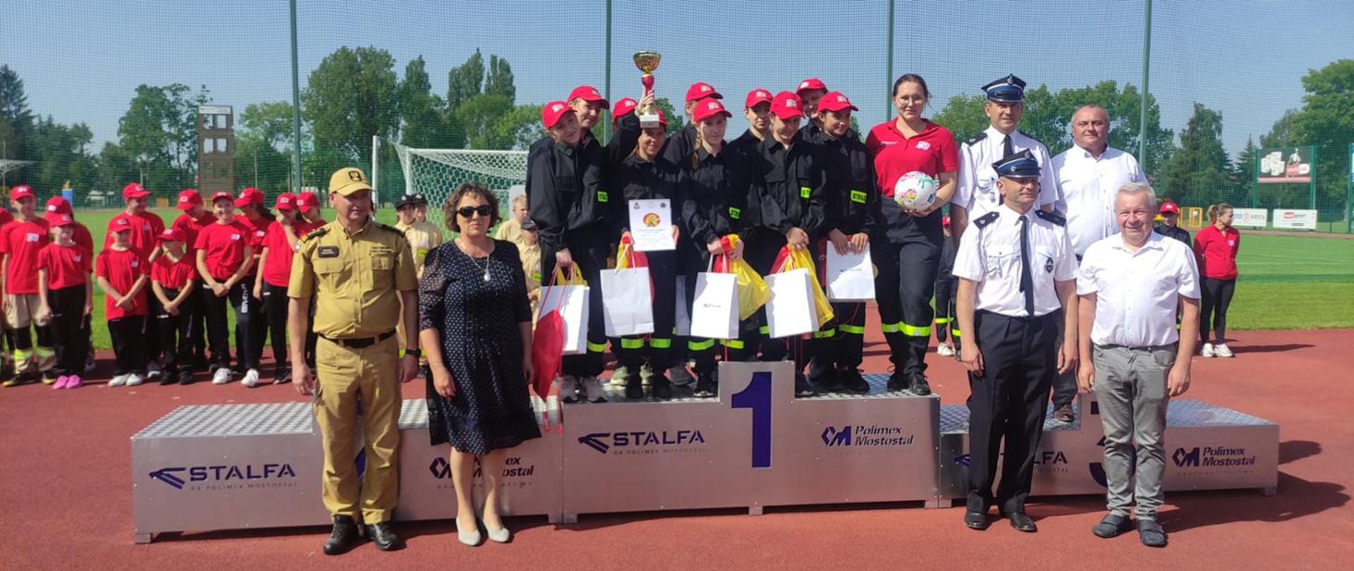 Zdjęcie grupowe na tle płyty głównej Stadionu Miejskiego w Sokołowie Podlaskim. metalowe podium, na którym stoi zwycięska drużyna dziewcząt. Obok podium organizatorzy zawodów, a w tle widzimy w szeregu młodzież uczestniczącą w zawodach.
