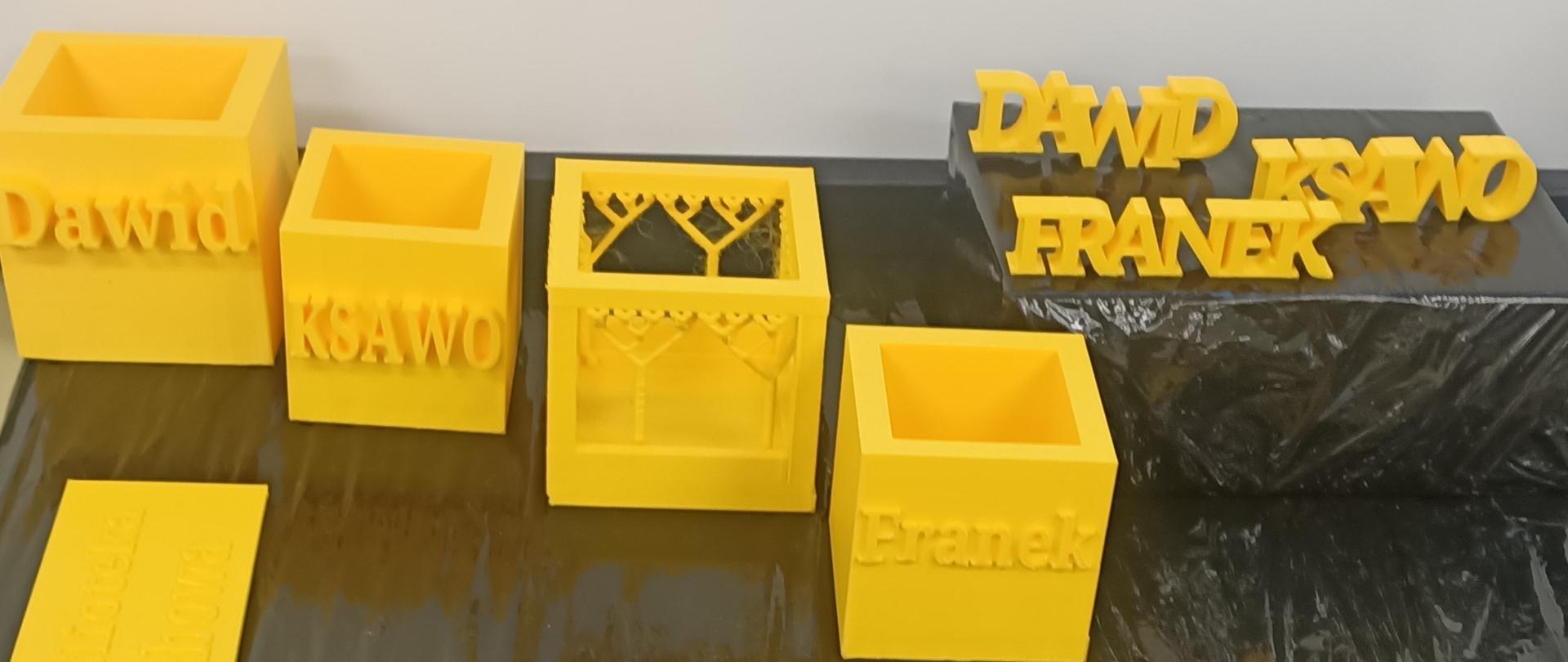 Na czarnej powierzchni stołu rozłożone są wydrukowane modele 3D- widnieją na nich napisy. Wszystkie wydruki są w kolorze żółtym.