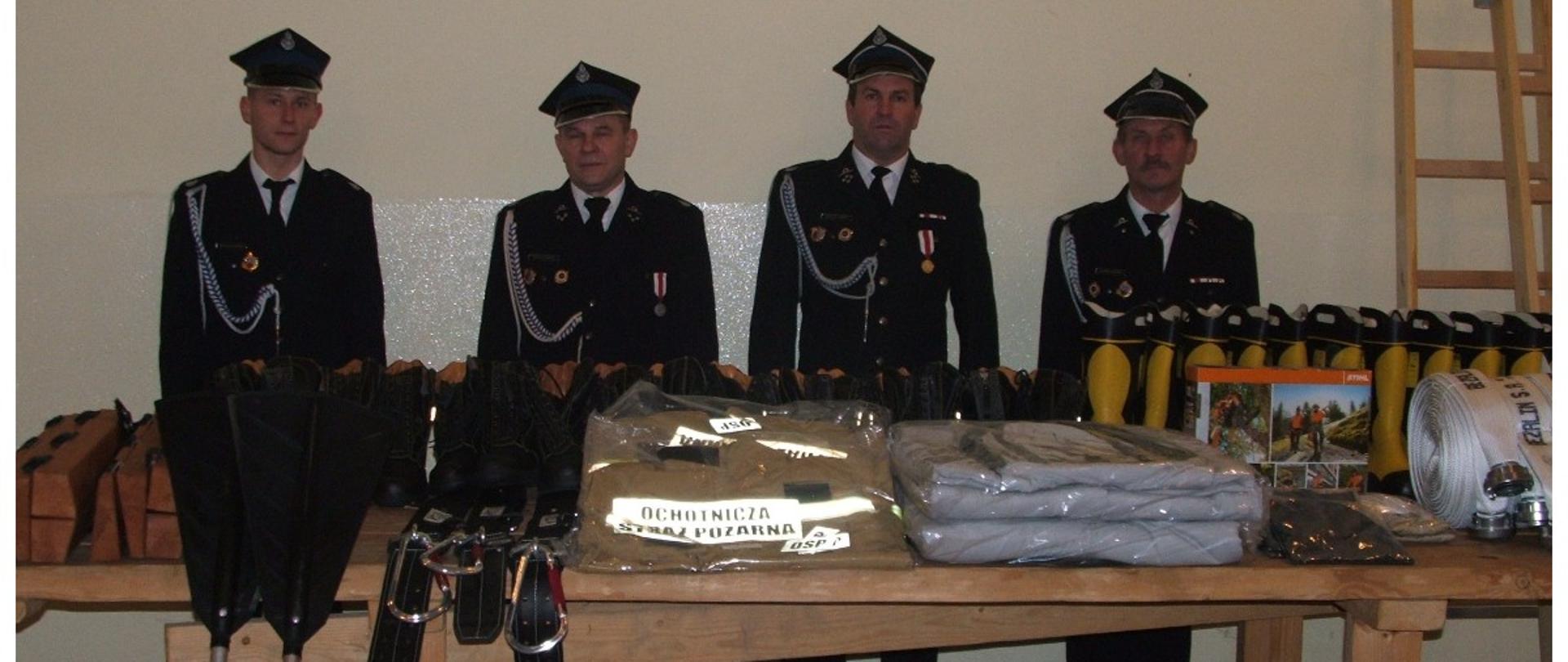 Zdjęcie przedstawia czterech umundurowanych strażaków pozujących do zdjęcia. Przed nimi jest stół, na którym prezentowany jest zakupiony sprzęt ratowniczo-gaśniczy. 