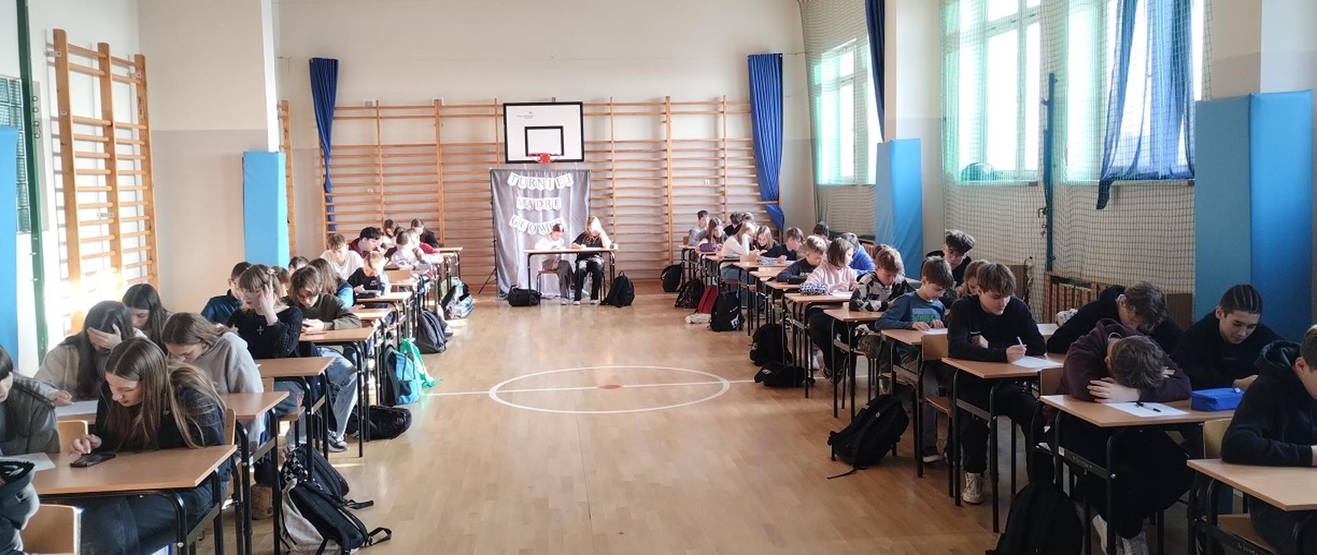 Młodzież siedząca i pisząca przy stolikach w dużej sali gimnastycznej .Stoliki ustawione przy ścianach sali. 