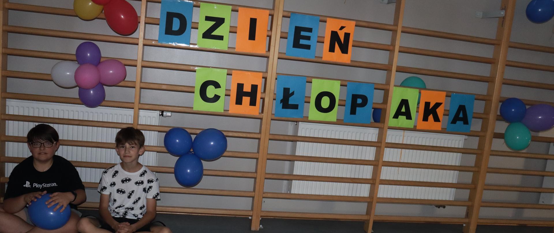 Dwóch chłopców siedzi na podłodze w sali gimnastycznej, na barierkach napis dzień chłopaka