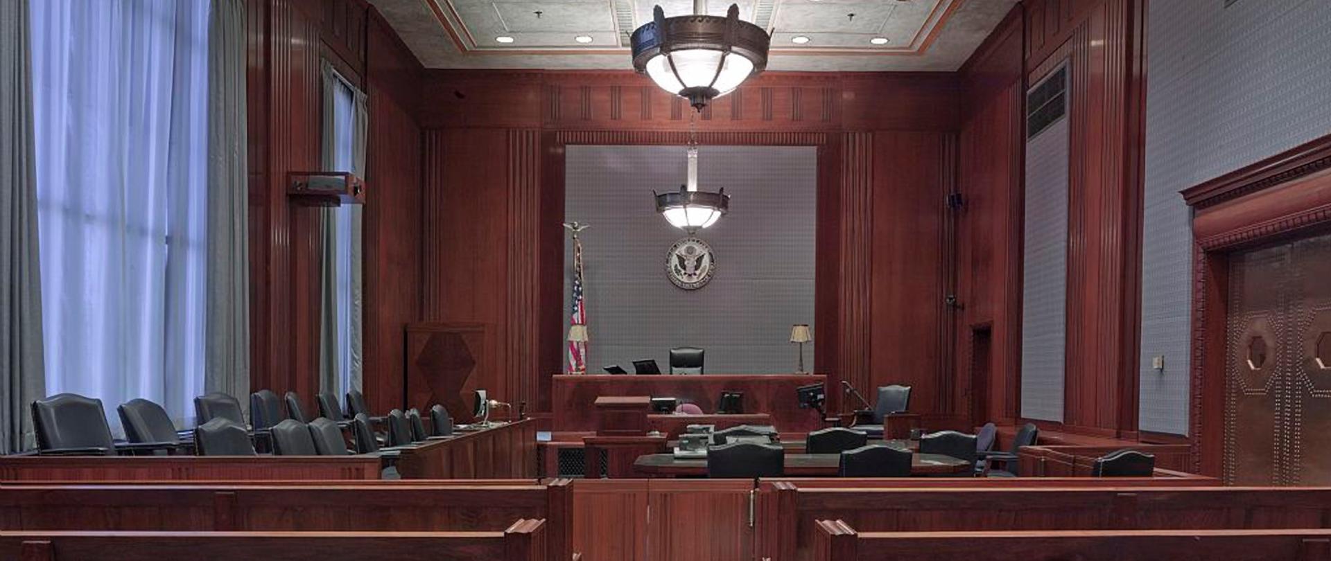Zdjęcie przedstawia wnętrze sali sądowej wewnątrz budynku. Na środku sufitu zwisa lampa, na pierwszym planie widać ławy a dalej w dalszej perspektywie krzesła.