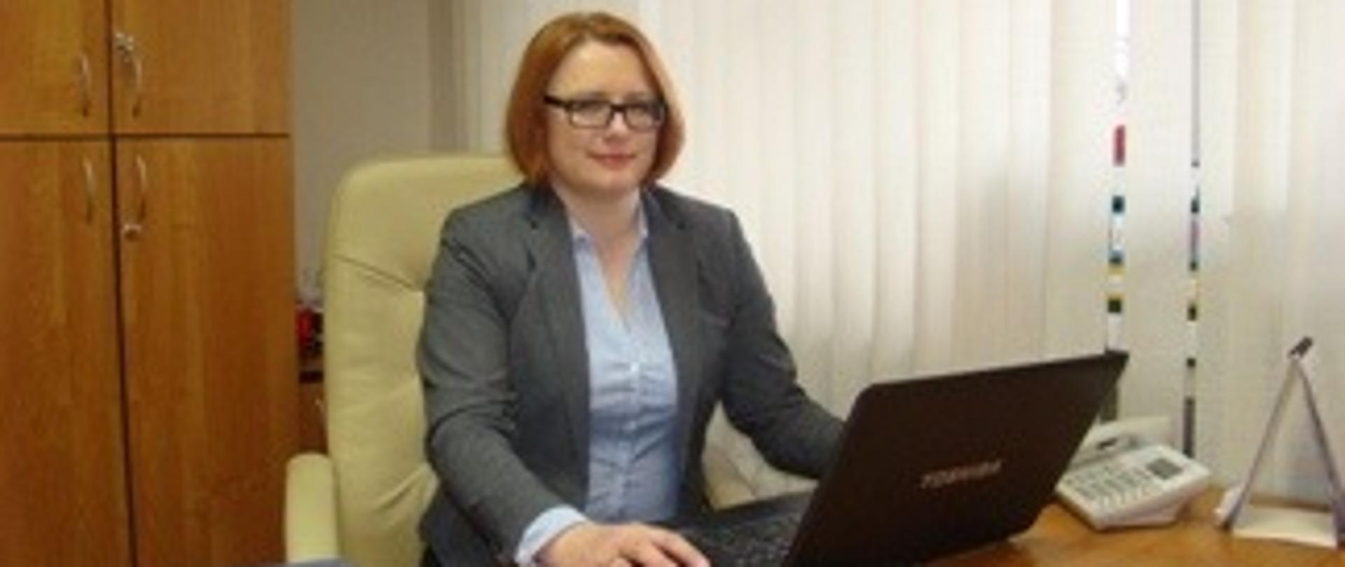 sekretarz siedzi za biurkiem przed laptopem w tle szafa po prawej stronie okno i żaluzje na biurku dokumenty widoczny telefon z prawej ubrana w popielatą marynarkę pod marynarką niebieska bluzeczka włosy w kolorze rudym