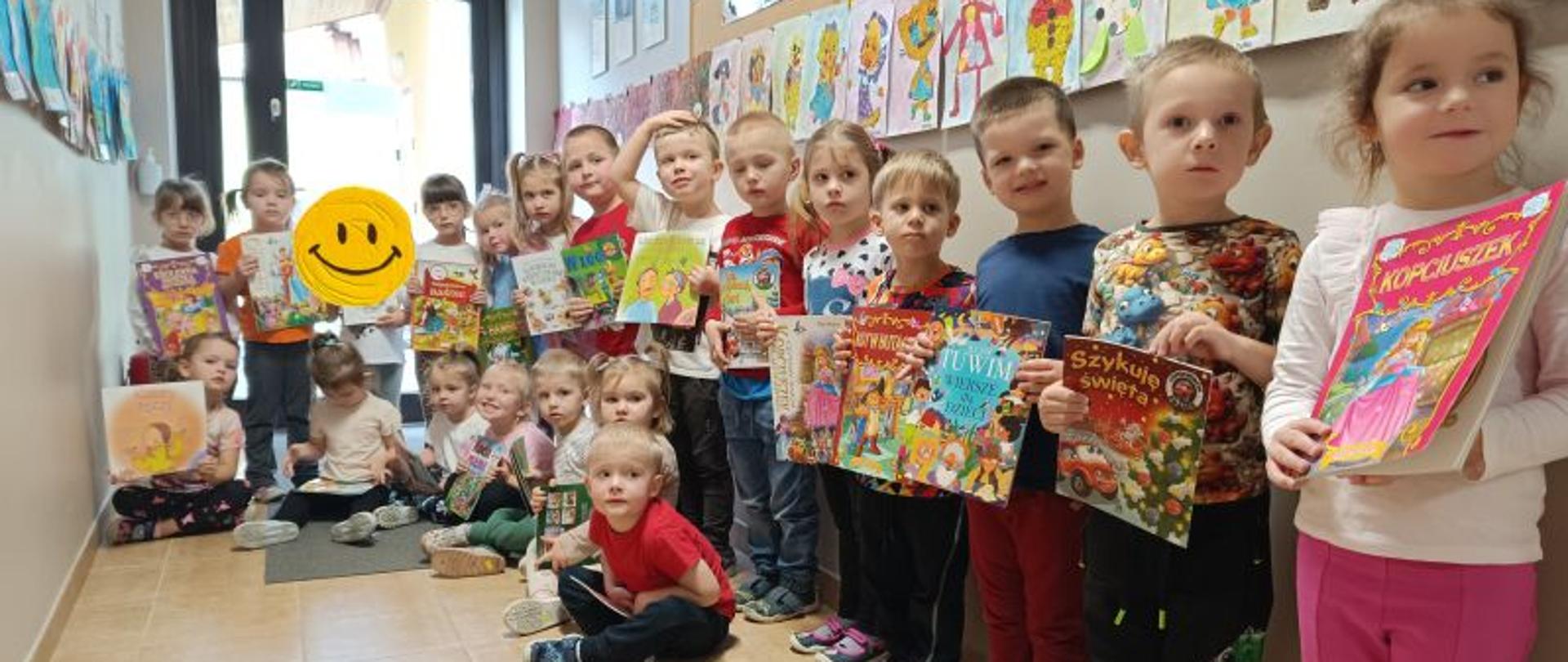 Dzieci z grupy Tygyrsków stoją i siedzą, w rękach trzymają książki. Za nimi powieszone prace plastyczne