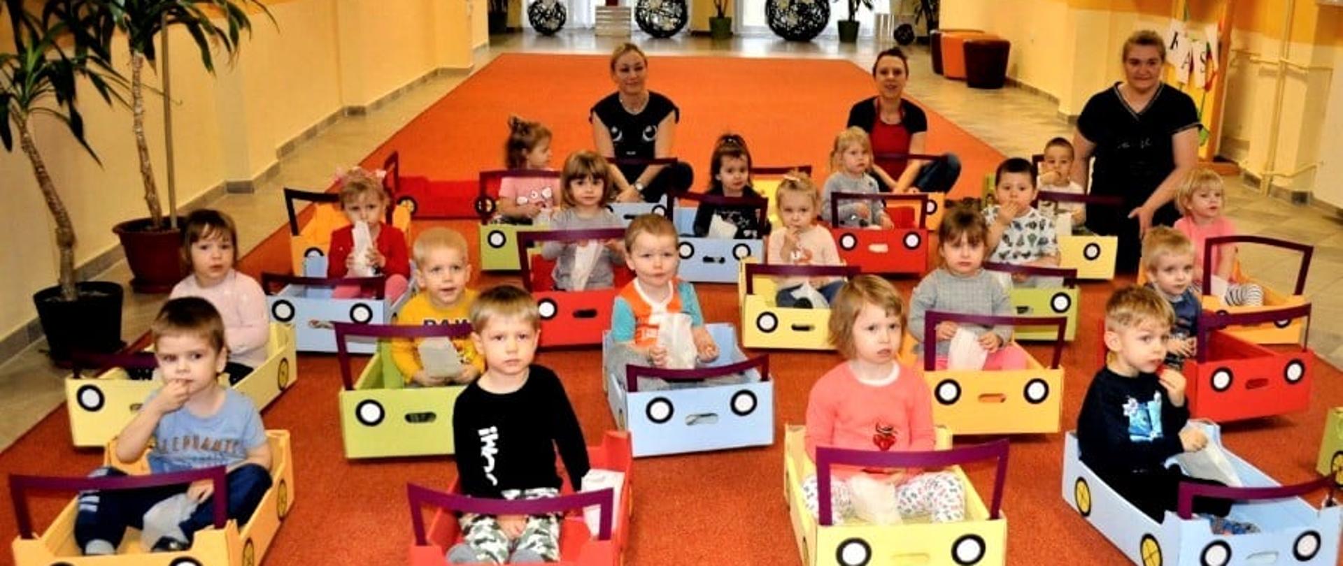 Kilkanaście osób (małych dzieci) siedzi w stworzonych z kartonu samochodach na podłodze. Za nimi, w tle, kucają trzy dorosłe osoby.