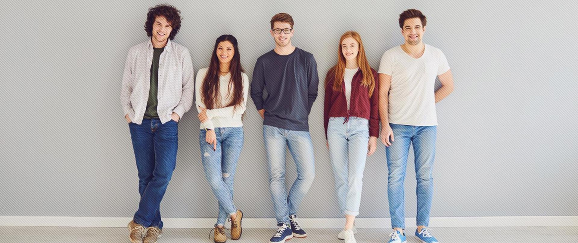 grupa pięciu osób stoją oparci o ścianę na zmianę mężczyzna kobieta mężczyzna kobieta i mężczyzna. Wszyscy w jeansach ubrani młodzieżowo. Dominuje kolor szary bo taka jest podłoga oraz ściana. 