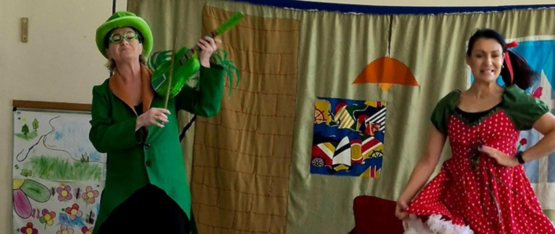 Dwójka aktorów w wyrazistych kostiumach odgrywa scenę przed grupą przedszkolaków siedzących na podłodze. Po lewej stronie artysta w zielonym surducie i kapeluszu pozoruje grę na zielonych skrzypcach, podczas gdy po prawej stronie aktorka w czerwonej sukience w grochy dynamicznie porusza się na tle materiałowej dekoracji.
