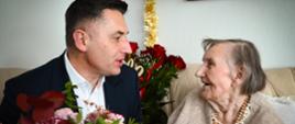 Przedstawiciel miasta rozmawia z seniorką siedzącą na kanapie, wręczając jej bukiet kwiatów w trakcie jubileuszowej wizyty.