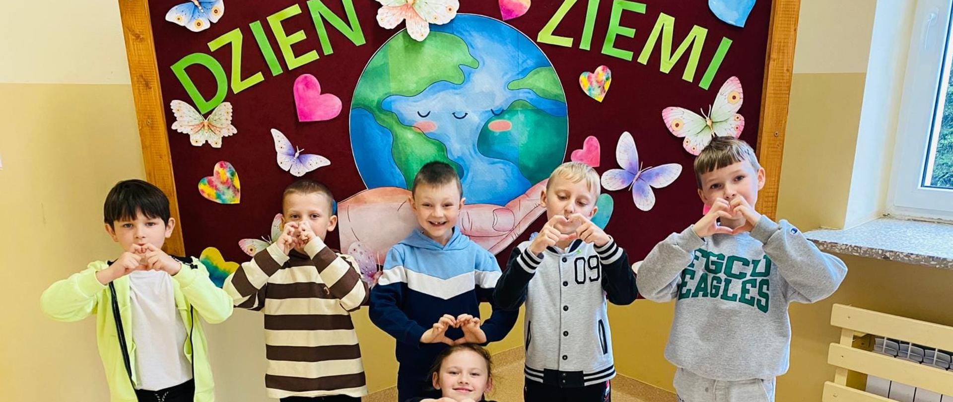Uczniowie klasy pierwszej czyli czterech chłopców i jedna dziewczynka stoją przed gazetką, na której widnieje napis "Dzień ziemi" i kula ziemska otoczona kwiatami i motylkami. Dzieci pokazują serduszko uformowane z palców dłoni