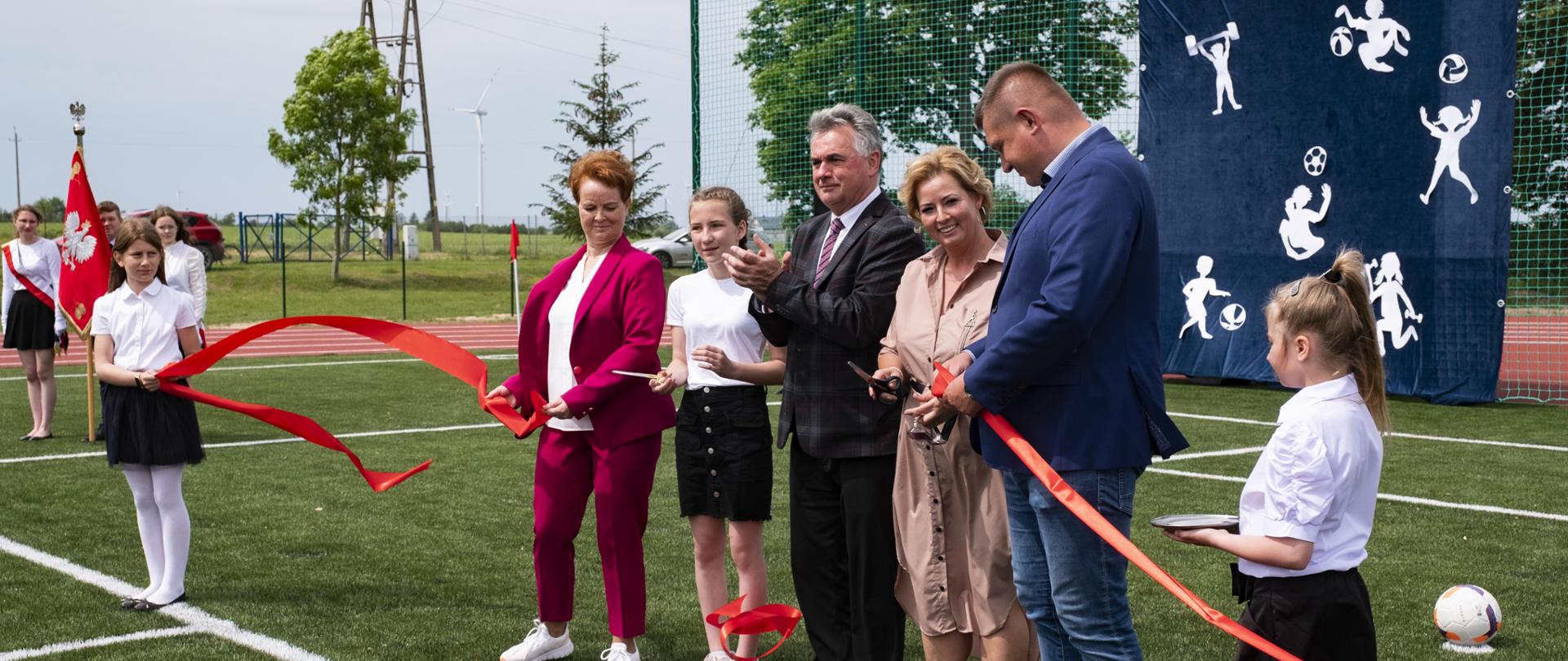 Burmistrz Janusz Piechocki, dyrektor szkoły w Lipinach Janina Szlufarska-Kłosek i 5 innych osób przecinają czerwoną wstęgę na boisku w Lipinach, w tle delegacja młodzieży ze sztandarem szkolnym, piłkochwyty z grafiką z granatowym tłem i białymi postaciami uprawiającymi różne rodzaje sportu