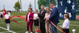 Burmistrz Janusz Piechocki, dyrektor szkoły w Lipinach Janina Szlufarska-Kłosek i 5 innych osób przecinają czerwoną wstęgę na boisku w Lipinach, w tle delegacja młodzieży ze sztandarem szkolnym, piłkochwyty z grafiką z granatowym tłem i białymi postaciami uprawiającymi różne rodzaje sportu