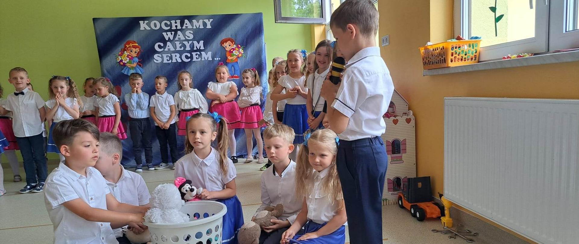 5 dzieci siedzi wokół plastikowego kosza z zabawkami. Jeden z chłopców mówi wiersz do mikrofonu. W tle widać pozostałe dzieci w odświętnych strojach. 