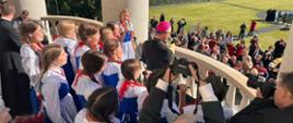 Biskup w czarnej szacie na balkonie wokół niego dzieci w biało niebieskich strojach spoglądają na ludzi stojących na trawniku