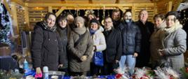 Zdjęcie przedstawia grupę kilkunastu uśmiechniętych osób pozujących do wspólnej fotografii wewnątrz drewnianej wiaty udekorowanej ciepłymi lampkami. Wszyscy są ubrani w zimowe kurtki i płaszcze, a jedna z kobiet ma na głowie opaskę z rogami renifera. Na pierwszym planie znajduje się długi stół przykryty niebieskim obrusem i zielonym tiulem, na którym wyeksponowano bożonarodzeniowe produkty: ozdobne słoiki, stroiki z igliwia oraz paczki z piernikami oznaczone cenami. W tle, po lewej stronie, widoczny jest termos gastronomiczny oraz akcesoria do serwowania ciepłych napojów.