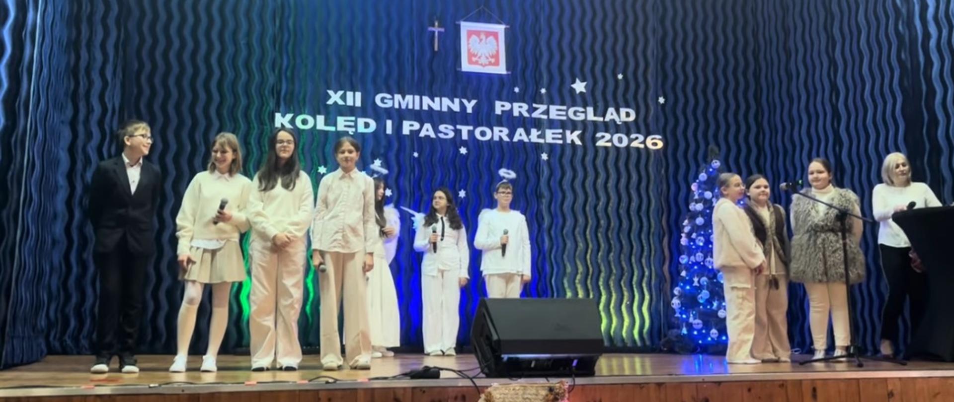 Dziesięcioro uczniów z klas 4-6 stoi na scenie. Siedmioro uczniów ubranych jest na biało. Jeden chłopiec ma na sobie czarny garnitur, a dwie dziewczyny przebrane są za pastuszków. Z tyłu widnieje biały napis na niebieskim tle: Dwunasty Gminny Przegląd Kolęd i Pastorałek 2026.