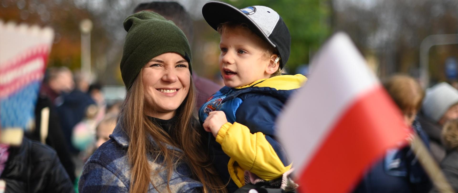 Na zdjęciu widzimy mieszkankę Konstantynowa, która trzyma swojego syna. Oboje mają szerokie uśmiechy na twarzach, są szczęśliwi i dumni. To piękny obraz miłości i więzi rodzinnej, który symbolizuje radość i dumę z przynależności do społeczności Konstantynowa.