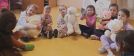 Przedszkolaki w swojej sali siedzą w kręgu w zabawę z przyniesionymi misiami, w tle tablica interaktywna z prezentacją związaną z misiem