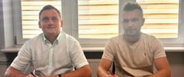Dwóch mężczyzn siedzi przy drewnianym stole w biurze na tle okna z roletami