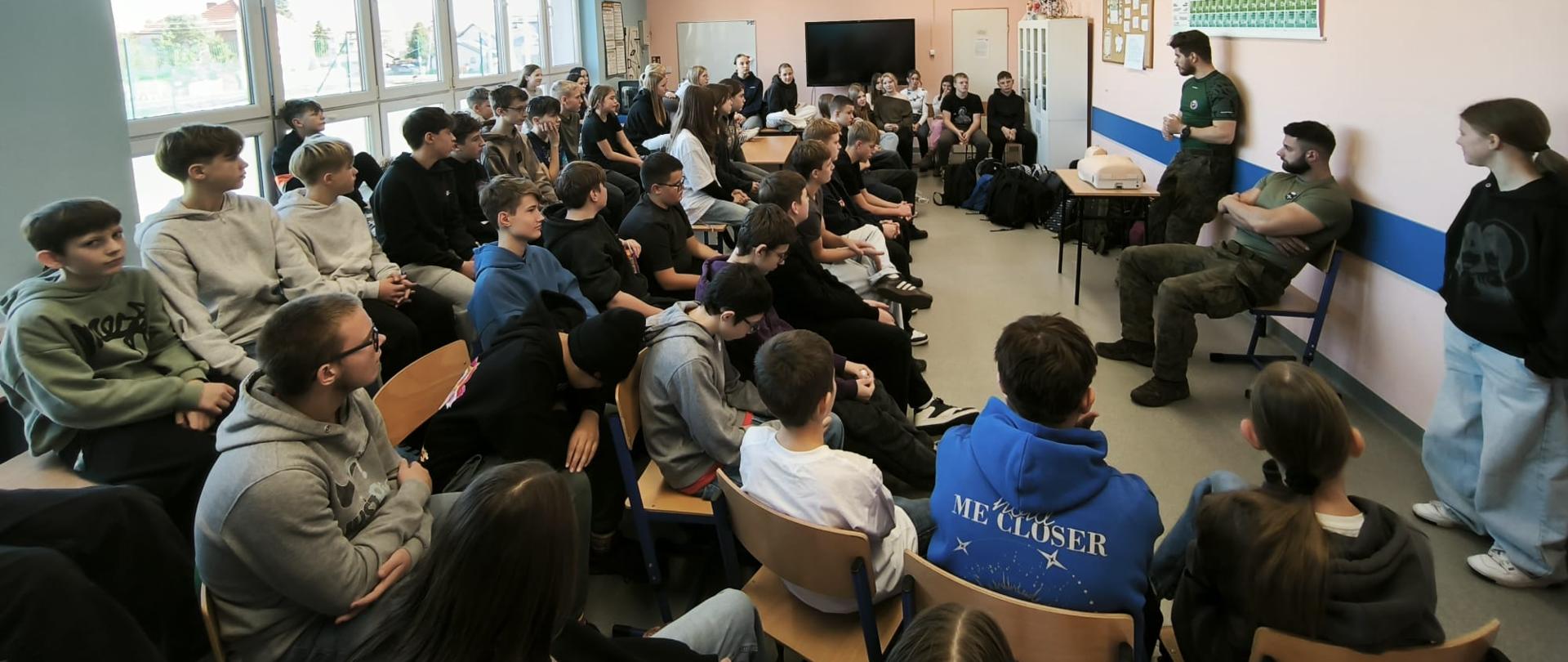 Uczniowie siedzą na krzesłach w sali i słuchają przemawiających żołnierzy.