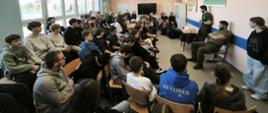 Uczniowie siedzą na krzesłach w sali i słuchają przemawiających żołnierzy.