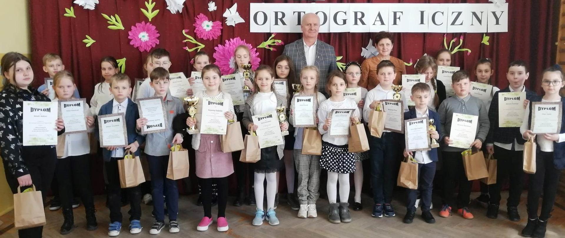 Na zdjęciu widzimy wszystkich biorących udział w konkursie ortograficznym, za nimi stoi przedstawiciel Gminy Szastarka Wójt Artur Jaskowski . Dzieci trzymają w ręce puchary, dyplomy, oraz siwe papierowe torebki z zawartością.