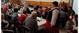 Grupa osób w różnym wieku siedząca przy stołach , rozmawia z Panią Sołtys ubraną w tradycyjny strój góralski. 