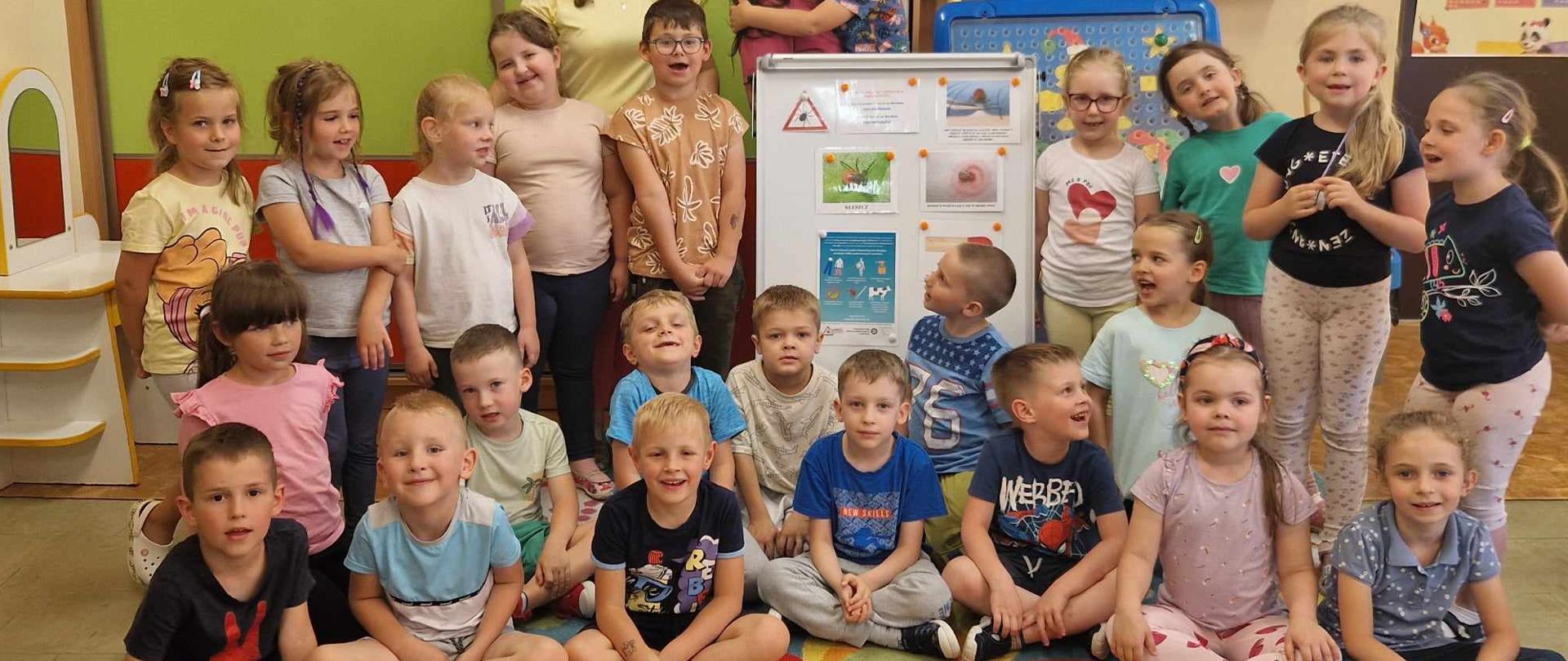 2 osoby dorosłe i grupa dzieci przy tablicy demonstracyjnej