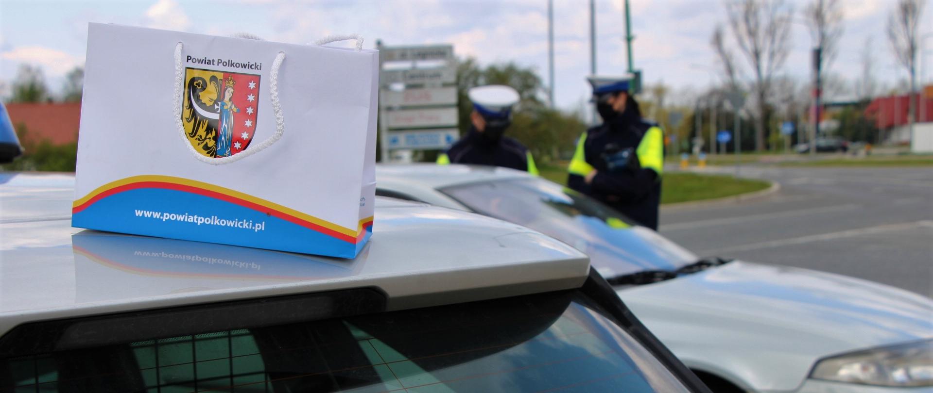 Dwa auta stojące na poboczu drogi. Na pierwszym aucie stoi torebka z herbem powiatu.