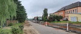 Rozbudowywany odcinek drogi, obsadzone nowe krawężniki, po prawej stronie znajduje się budynek szkoły. W oddali widać koparkę i samochody osobowe.