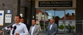 4 osoby stojące przed wejściem do szpitala powiatowego w Chodzieży, jedna przemawia do mikrofonów