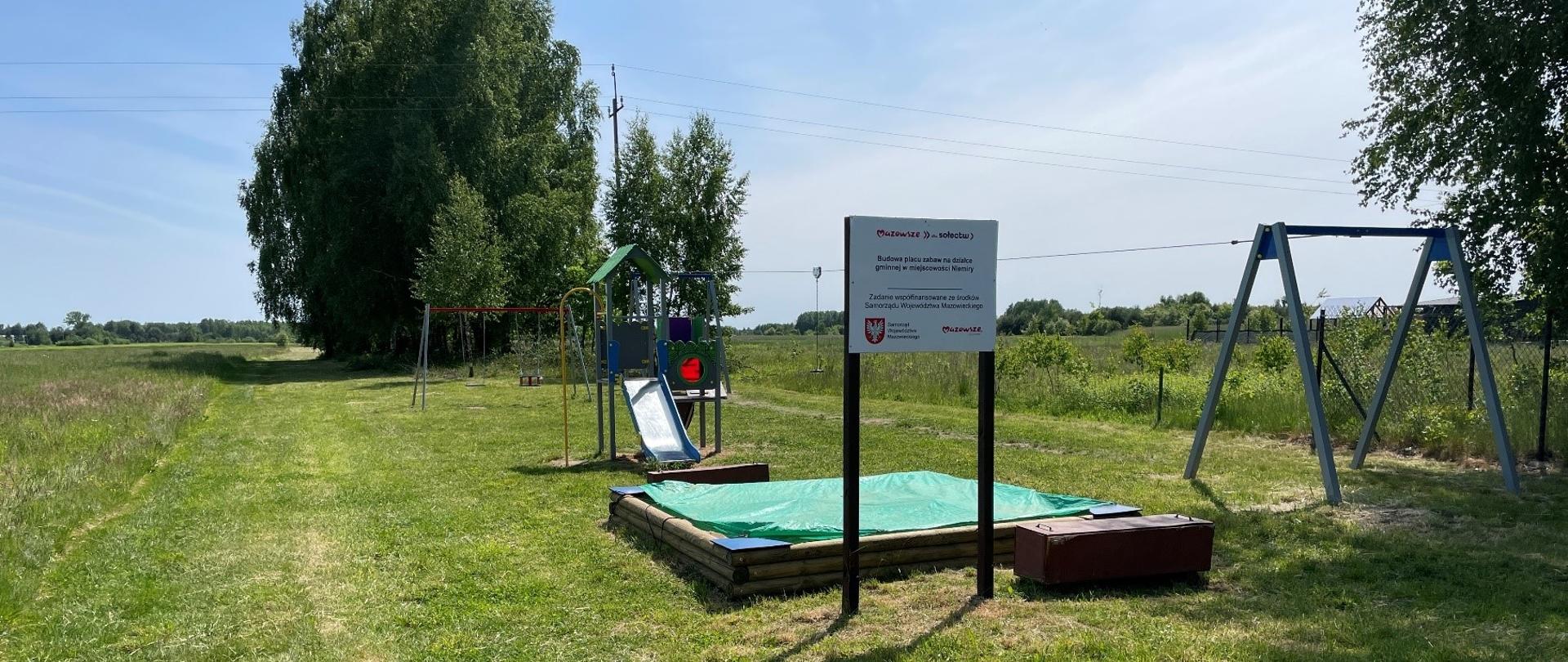 Plac zabaw na którym znajdują się: piaskownica zjeżdżalnia, huśtawka, zjeżdżalnia linowa oraz tablica informacyjna dot. finansowania.