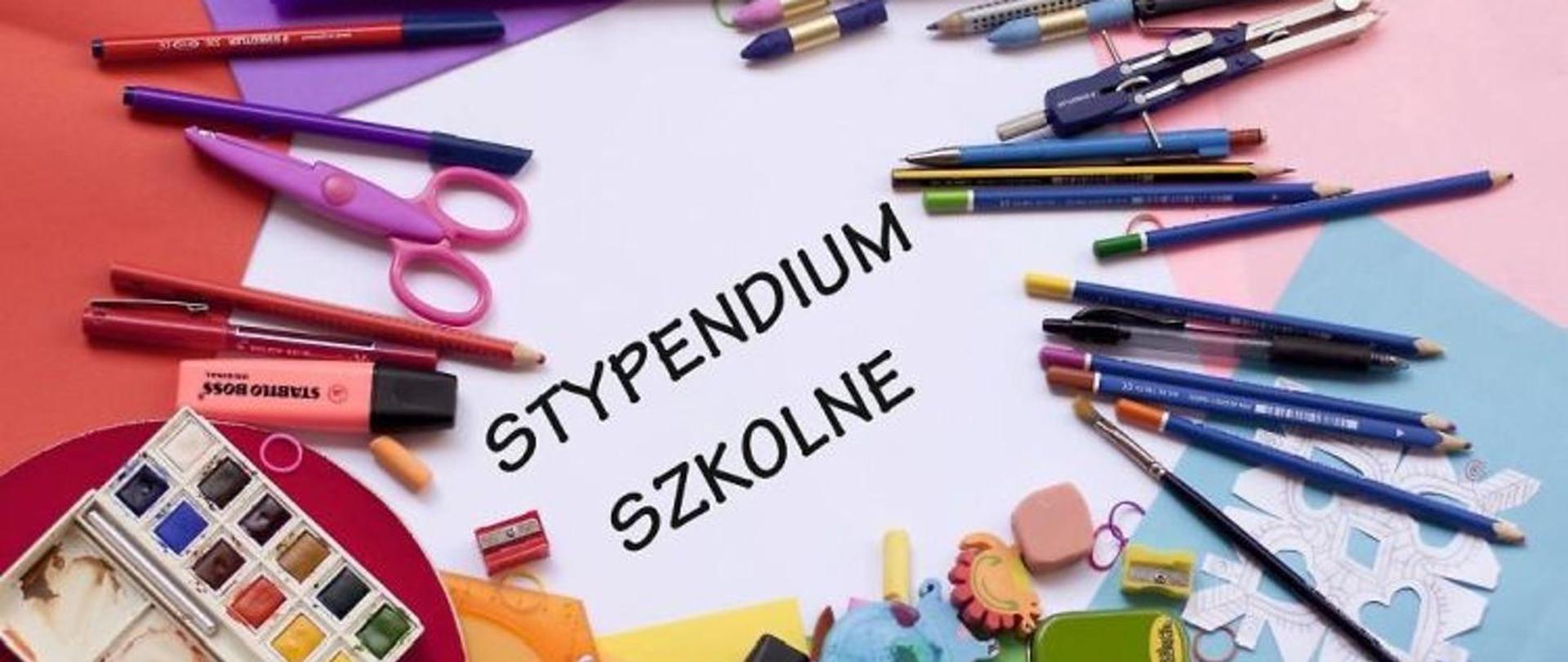 Materiały szkolne (pisaki, ołówki, długopisy itp) ułożone na kolorowych kartkach papieru. Na środku napis stypendium szkolne