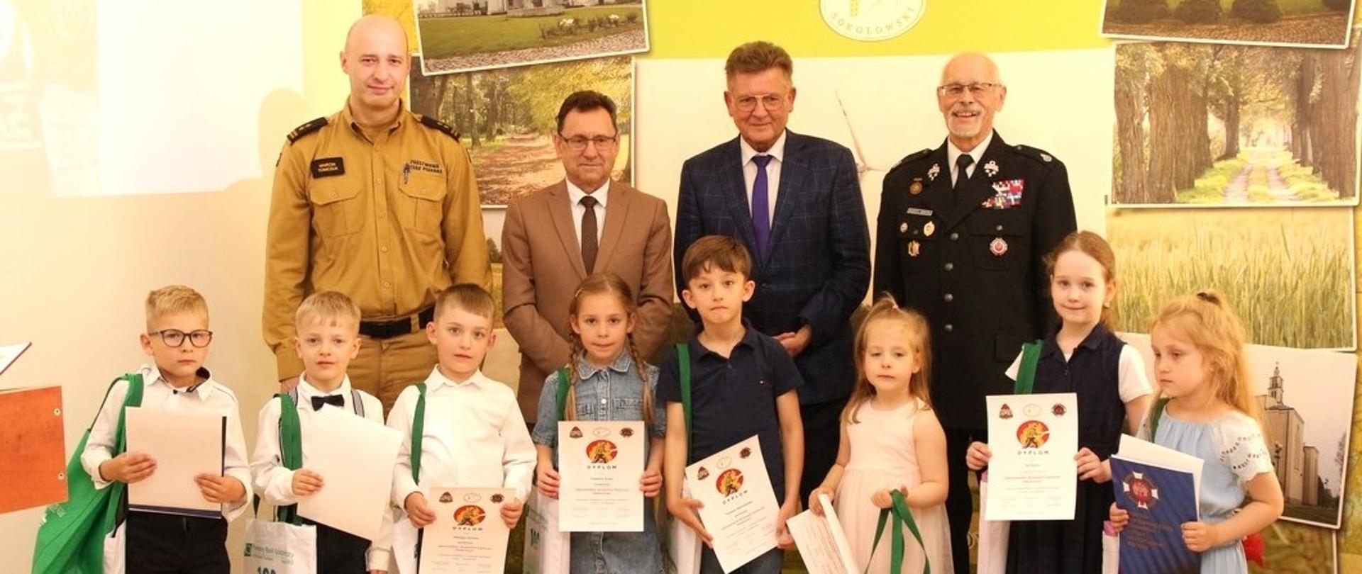 Zdjęcie grupowe wykonane w sali konferencyjnej Starostwa Powiatowego w Sokołowie Podlaskim podczas rozstrzygnięcia Konkursu Plastycznego ”Ziemia, Woda, Powietrze – strażacy na straży ochrony środowiska”. N zdjęciu laureaci konkursu trzymają dyplomy oraz nagrody w zielonych torebkach powiatowych, za nimi od lewej komendant PSP w Sokołowie Podlaski, starosta, prezes zarządu ZOSP RP w Sokołowie Podlaskim oraz przewodniczący rady powiatu. W tle zielona ścianka powiatu sokołowskiego, na której jest logo powiatu oraz zdjęcia zabytków z powiatu.
