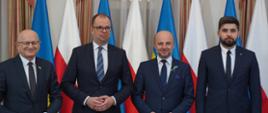Zdjęcie przedstawia cztery osoby stojące wewnątrz budynku na tle biało-czerwonych flag