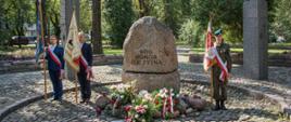 Widok ogólny na eliptyczny głaz pęknięty poprzecznie na pół z napisem Bóg, Honor Ojczyzna stanowiący centralną część pomnika Czynu Niepodległości w Sokołowie Podlaskim. Po obu stronach głazu widoczne sztandary szkolne, a pod nim złożone kwiaty i zapalone znicze.