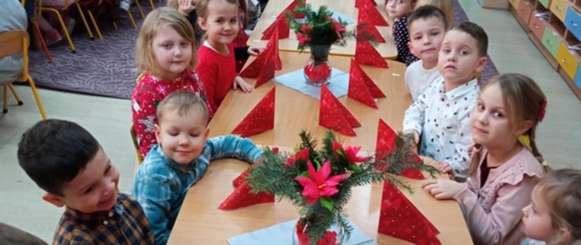 Przy stołach przystrojonych bożonarodzeniowymi dekoracjami siedzą przedszkolaki. Spoglądają w kierunku obiektywu aparatu i uśmiechają się.