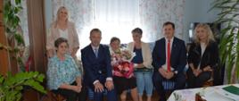 Zdjęcie przedstawia siedem osób pozujących do wspólnej fotografii w przytulnym, domowym wnętrzu. Sześć osób siedzi na kanapie i krzesłach, a jedna kobieta stoi z tyłu. W centrum znajduje się starsza kobieta trzymająca bukiety kwiatów oraz zieloną teczkę. Towarzyszą jej dwaj mężczyźni ubrani w eleganckie garnitury i trzy kobiety w jasnych żakietach oraz wzorzystej bluzce. W pomieszczeniu widoczne są kwiatowe zasłony, rośliny doniczkowe, telewizor i stół nakryty obrusem. Uczestnicy spotkania uśmiechają się do obiektywu. Fotografia przedstawia uroczyste, serdeczne spotkanie w kameralnej atmosferze.