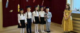 5 uczniów klasy 1 odświętnie ubranych przedstawia program artystyczny na szkolnym korytarzu, po prawej stronie stoi uczennica klasy 3 w żółtej sukni i koronie na głowie, za dziećmi na ścianie bordowe płótno z napisem "Pasowanie na ucznia" i papierową sylwetą sowy