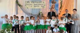 Grupa dzieci w białych bluzeczkach i jasnozielonych elementach strojów (spódniczki, muchy, szelki) pozuje na scenie z dwiema opiekunkami i dwoma mężczyznami. W centrum widoczny jest dyplom dla grupy „Żabki”.