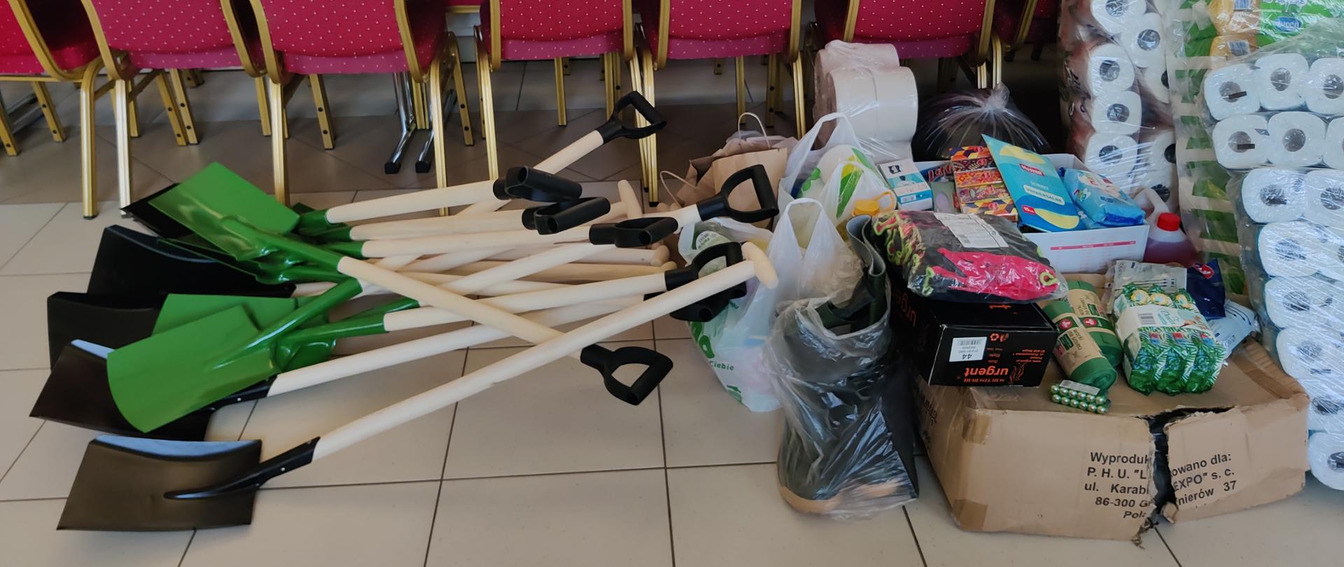 Łopaty i zielone szpadle leżące na podłodze, za nimi czerwone krzesła i stoły z artykułami spożywczymi, obok na podłodze kartony i środki czystości oraz duże paczki papieru toaletowego