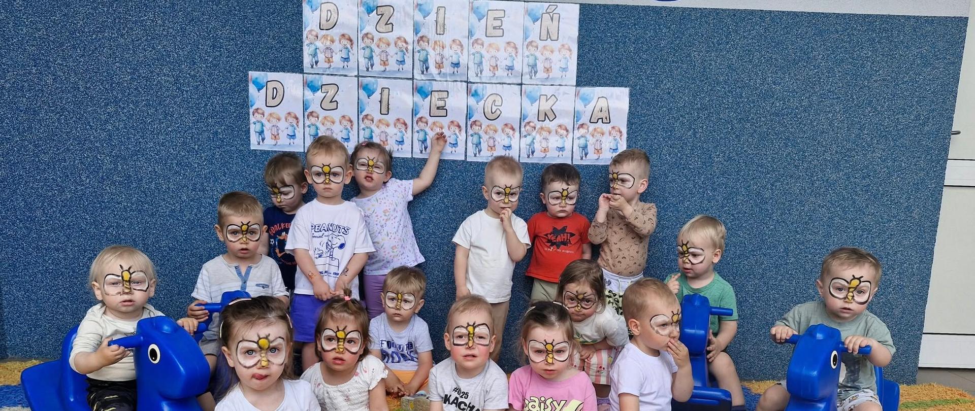 Dzieci z grupy Pszczółki świętujące Dzień Dziecka na tarasie. Wszystkie mają namalowane na twarzach pszczółki, pozują do zdjęcia w tle napis Dzień Dziecka.
