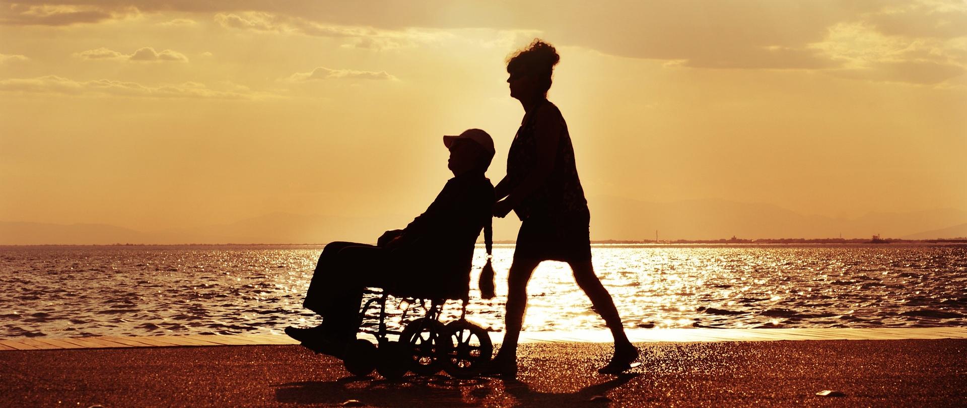 Zdjęcie kobiety pchającej wózek inwalidzki z siedzącym w nim starszym mężczyzną, w tle widok z plaży na zatokę i zachód słońca