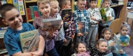 Uczniowie klasy pierwszej po złożeniu uroczystego przyrzeczenia dotyczącego poszanowania książek pozują do zdjęcia na tle półek z książkami, w rękach trzymają wypożyczone książki